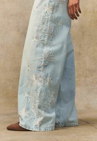 Jeans de mezclilla bordados en azul claro con patrones florales a lo largo de los lados, que presentan un diseño de pierna ancha y un dobladillo sin terminar. Zapatos marrones de puntera afilada visibles.