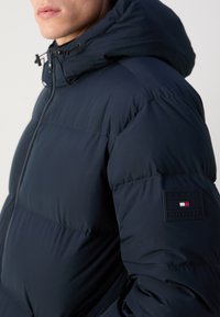 Man wearing navy blue puffer jacket with hood, visible Tommy Hilfiger logo patch on upper sleeve, zipped up to neck.