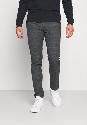 Mand iført slim-fit gråternet bukser og hvide sneakers, gående mod en ensfarvet lys baggrund.