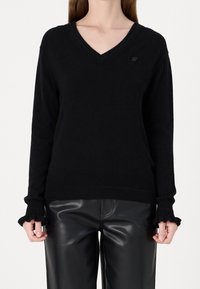 Pull noir en V en tissu doux, avec des manches longues à volants et un logo discret sur la poitrine. Associé à un pantalon noir.