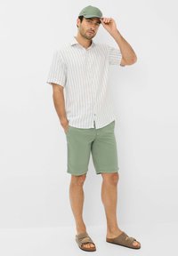 Camicia a maniche lunghe con un leggero motivo a righe, pantaloni corti verdi e un cappellino verde. Sandali neutri completano l'outfit informale.