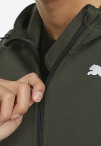 Olijfgroene zip-up jack van zacht materiaal, met een zwarte rits en een klein wit logo op de borst. Gladde textuur, ontworpen met een capuchon.