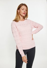 Maglione a maglia rosa chiaro con motivi testurizzati e piccole aperture, colletto rotondo e maniche lunghe. Abbinato a pantaloni neri.