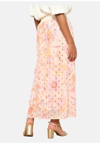 Jupe longue rose avec des motifs floraux et des pois dorés, fabriquée en tissu léger. Associée à des sandales à talons dorées.