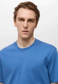 Tricou din bumbac albastru, cu guler rotund și maneci scurte. Materialul pare neted, cu un ușor luciu și are o croială regulată.