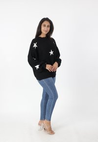 Zwarte gebreide trui met witte sterrenpatronen, gecombineerd met blauwe skinny jeans en transparante haksandalen. Eenvoudige, ontspannen pasvorm.