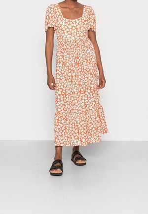 Female model wearing an orange floral midi dress with puff sleeves and black sandals, standing on a plain light gray floor and wall.