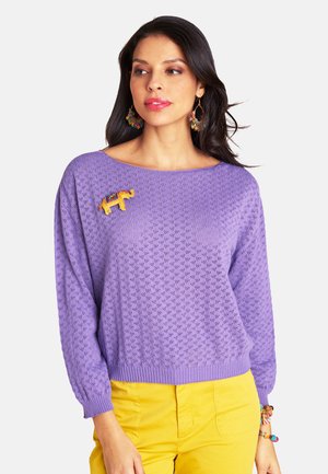 Frau trägt lavendelfarbenen Strickpullover mit kleinem Elefanten-Brosche, gelbe Hose, bunte Creolen und Perlenarmband, schaut nach rechts.