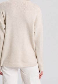 Un pull en laine beige avec un motif texturé porté par une personne vue de dos, associé à un pantalon blanc cassé sur un fond neutre.