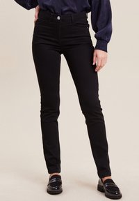 Jean stretch noir taille haute, coupe slim, poches avant et arrière, et un bouton argenté brillant. Associé à des mocassins noirs brillants.