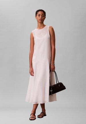 Kvinde iført en hvid midikjole uden ærmer, der holder en sort håndtaske, kombineret med sorte flade sandaler, stående mod en grå baggrund.