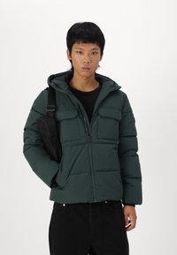 Dark green puffer jacket with a zipper and two chest pockets. Features a hood, quilted texture, and is paired with a black shoulder bag.