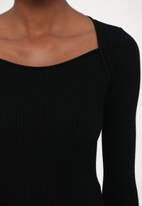 Svart ribbet topp med firkantet hals, lange ermer og en tettsittende design, med et teksturert stoff for ekstra dimensjon.