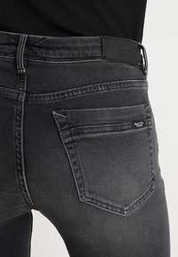 Jeans de mezclilla negros con corte ajustado, que presentan una etiqueta de marca de cuero en la cintura y dos bolsillos traseros con costuras decorativas.