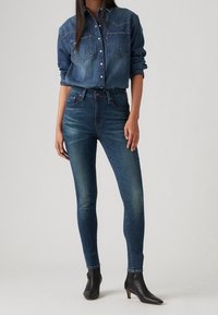 Camisa de denim en azul oscuro con dos bolsillos en el pecho, diseño de botones y mangas arremangadas. Jeans skinny azul oscuro con un ligero desvanecimiento. Botines negros hasta el tobillo.