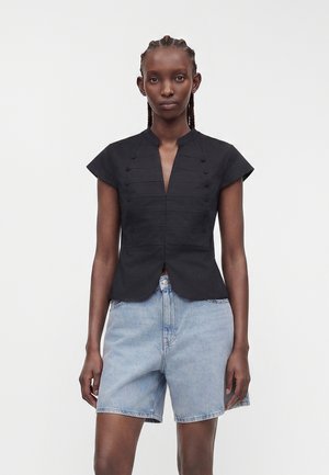 Femme aux cheveux tressés porte un chemisier noir ajusté à manches courtes avec des boutons et un short en jean bleu clair, face à un fond blanc.