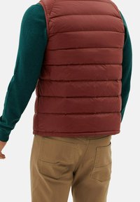 Bordeauxrote Steppweste mit gestepptem Design; getragen über einem türkisfarbenen Langarmshirt. Sichtbare beigen Hosen mit Gesäßtaschen.