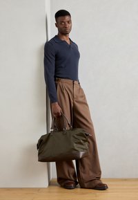 Olive green duffel bag with dark brown leather accents, textured fabric, and sturdy handles, resting on wooden floor beside a person.
