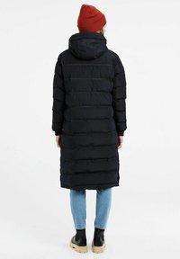 Sort polstret frakke med hætte, der har vandrette sømme og en lang pasform. Sammen med blå jeans og sorte klodsede støvler. Rød hue på hovedet.