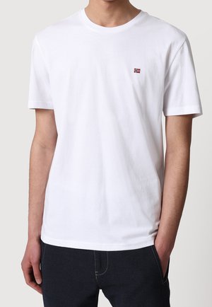 White cotton t-shirt with short sleeves, round neckline, and small red and blue flag emblem on the left chest. Smooth texture.
