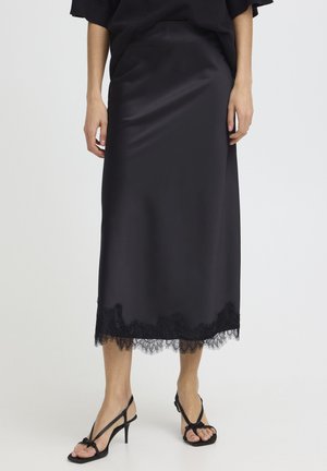 Žena v čiernej saténovej midi sukni s čipkovaným lemom a čiernych sandáloch na vysokom podpätku so šnurovaním, stojaca na jednoduchom pozadí.