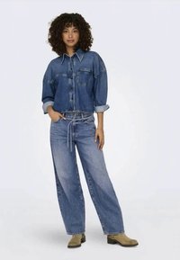 Jeans overshirt met zakken en opgerolde mouwen, gecombineerd met wijde spijkerbroeken, beide in lichtblauw, aangevuld met beige laarzen.