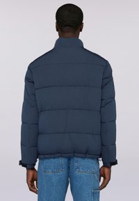 Uomo che indossa una giacca imbottita blu scuro e jeans blu, in piedi con la schiena rivolta verso la telecamera su uno sfondo chiaro e uniforme.