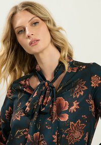 Donker bloemenblouse van een soepele stof met een striknek. Kleuren zijn diep groen, oranje en bordeauxrood met ingewikkelde bloempatronen.