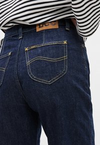 Tumesinised teksapüksid kollase õmblusega, millel on nahast logomärk ja kaks tagataskut kõverate õmblustega. Vöökohast ülespoole paistab triibuline särk.