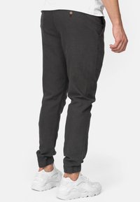 Donkergrijze joggingbroek met elastische boorden, zijzakken en een knoopdetail op de rug, gedragen met witte sportschoenen. Zachte stoftextuur.