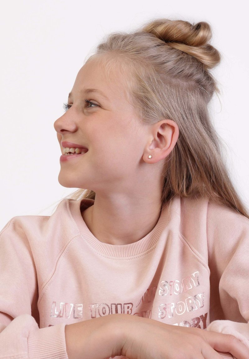 Lichtroze sweatshirt met glanzende tekst, voorzien van een ronde halslijn en raglanmouwen. Lange blonde haren in een half-opgestoken stijl, kleine gouden oorbellen.