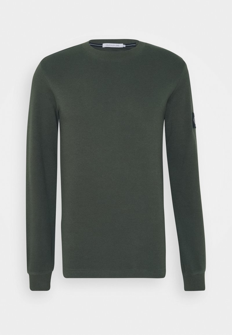 Dark green long-sleeve sweater in a ribbed texture with a round neckline. Features a logo patch on the sleeve.