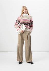 Donna che indossa un maglione a righe rosa, beige e marrone, pantaloni beige a gamba larga, scarpe nere, con in mano una borsa bianca, in piedi contro uno sfondo bianco.