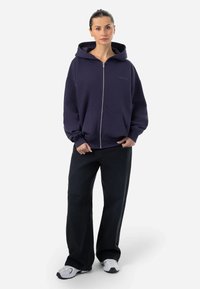 Mornarska zip-up hoodie sa velikom kapuljačom, kengurskom džepu i rebrastim rubom. Crne široke hlače s istrošenim detaljima. Bijele tenisice.