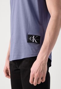 Purple cotton T-shirt with a crew neckline, short sleeves, and a black label featuring "Calvin Klein Jeans" and a white logo on the side.