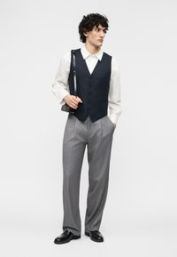 Νέος άνδρας που φοράει λευκό πουκάμισο, μαύρο γιλέκο, γκρι παντελόνι, μαύρα παπούτσια, κρατώντας μια μαύρη τσάντα ώμου, στέκεται μπροστά σε απλό φόντο.