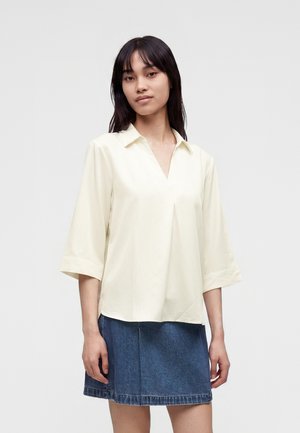 Femme aux cheveux foncés portant un chemisier crème à col en V avec col et manches trois-quarts, associé à une jupe en jean bleue.