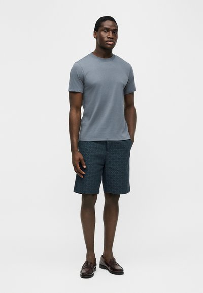 Άνδρας που στέκεται φορώντας γκρι κοντομάνικη μπλούζα, σκούρα σορτς με σχέδια και σκούρα καφέ μοκασίνια μπροστά σε απλό φωτεινό φόντο.