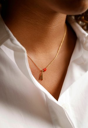 Collier en chaîne dorée avec un petit cœur rouge et un pendentif en forme de lettre "A", porté sur une chemise blanche à col.