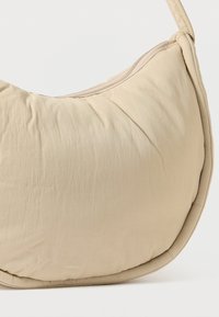 Μπεζ τσάντα ώμου με απαλό, καπιτονέ υφή, καμπυλωτό σχήμα και φερμουάρ κατά μήκος του πάνω μέρους. Διαθέτει διακριτική σήμανση.