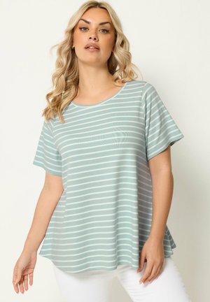 Kvinde iført en lyseblå og hvid horisontalt striped kortærmet løstsiddende skjorte og hvide bukser mod en ensfarvet baggrund.
