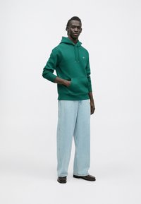 Grøn hættetrøje lavet af blødt materiale med en kængurulomme og justerbar snøre, kombineret med lyseblå løse jeans og sorte sko.