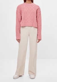 Suéter corto de punto trenzado en color rosa con mangas largas, combinado con pantalones de pierna ancha en un tono beige claro y zapatillas blancas.