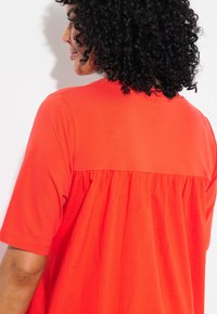 Camicia di cotone rossa con maniche corte, presenta un design raccolto sulla schiena e una texture morbida. Il colletto è di tipo crew senza decorazioni.