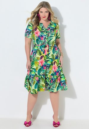 Nainen pukeutuneena vihreään, kukalliseen nappikiinnitteiseen mekkoon, jossa on lyhyet hihat, ja pinkkeihin korkokenkiin, joissa on rusetit, poseeraamassa valkoista taustaa vasten.