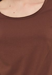 Κοντινό πλάνο στο ανώτερο μέρος του στήθους και τη γραμμή του λαιμού ενός ατόμου που φοράει καφέ αμάνικο τοπ με ορατές σπίλους στο δέρμα.