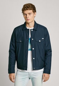 Chaqueta ligera de color azul marino, con cierre de botones en la parte delantera, dos bolsillos en el pecho y un cuello plano, usada sobre una camiseta gráfica blanca.