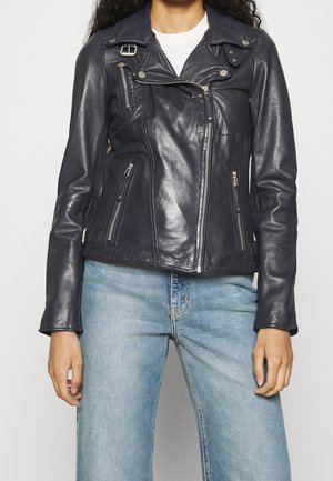 Personne portant une veste en cuir noire de motard avec des zips et un jean bleu clair sur un fond uni.