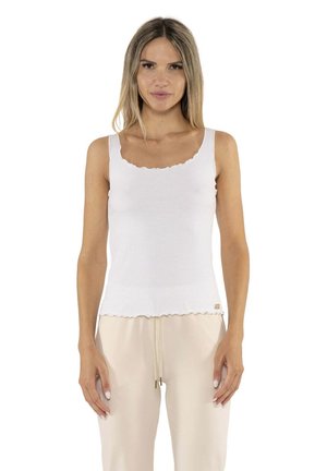 Une femme porte un haut blanc sans manches aux bords festonnés et un pantalon beige clair à cordon de serrage, sur un fond blanc uni.