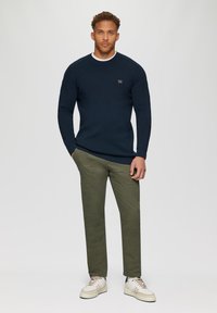 Marineblauwe gebreide trui met een geribbelde textuur en witte kraag, gecombineerd met olijfgroene broek en crèmekleurige sneakers, met een klein logo-detail.